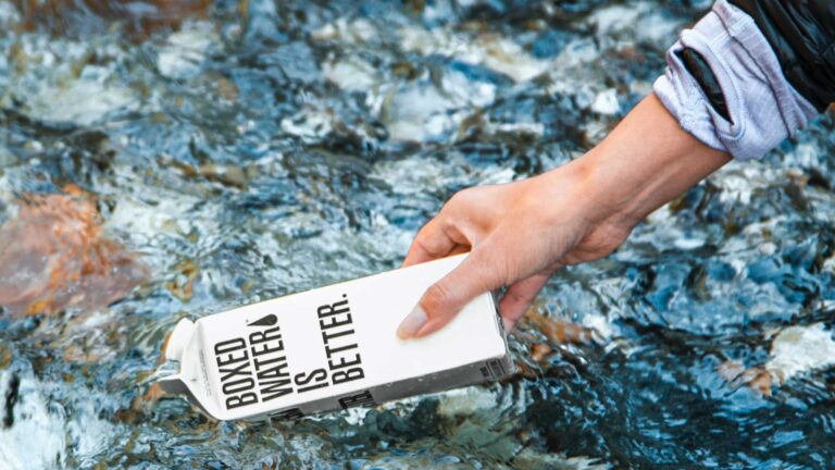 Photo of a woman's hand holding a boxed liquid container that has the words "Boxed water is better" on it