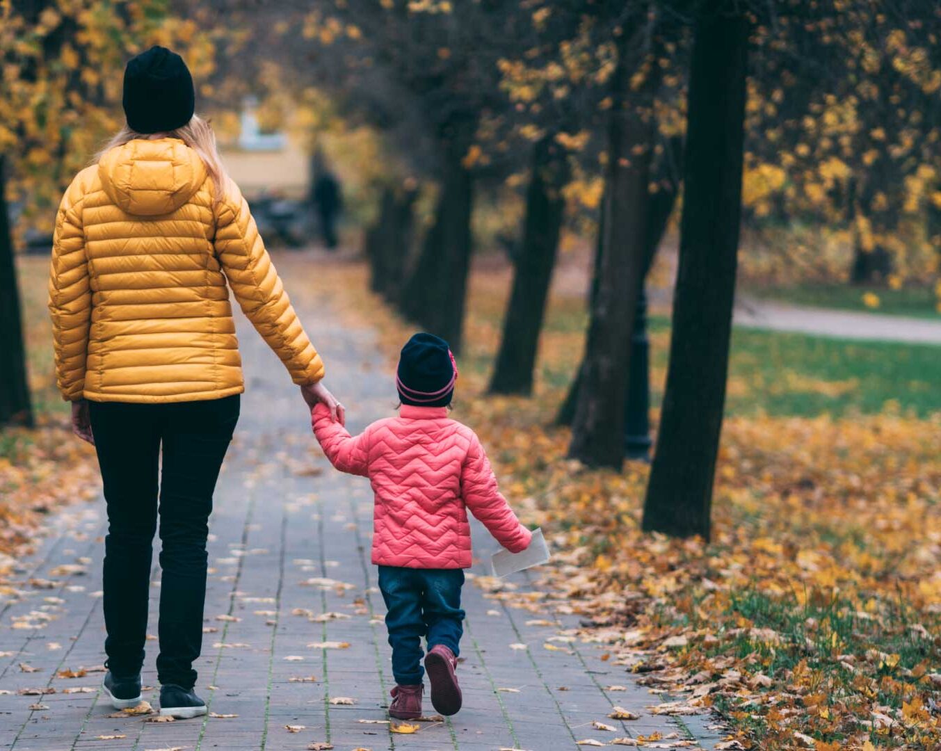 A photo of a woman and child walking, taken from behind. The woman is holding the child's hand. Trees and fall leaves line the path ahead of them. Credit Krzysztof Kowalik.