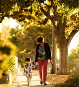 Photo of a woman and child walking on a sidewalk, with a bush on one side and trees in the background on the other. The woman and child are holding hands. Photo by Sue Zeng on Unsplash