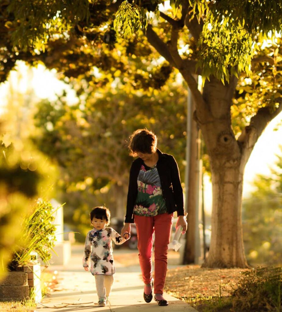 Photo of a woman and child walking on a sidewalk, with a bush on one side and trees in the background on the other. The woman and child are holding hands. Photo by Sue Zeng on Unsplash