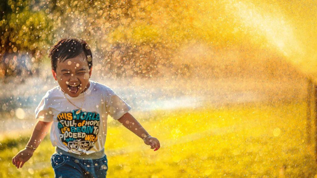 Photo of a boy running through a water sprinkler. Sunlight filters through that water from behind him. Credit Mi Pham.