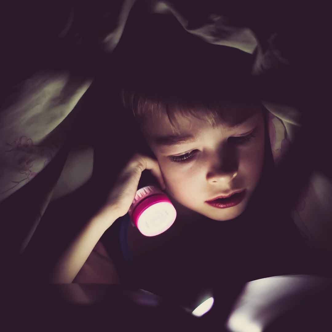 Photo of a child reading with a flashlight under his blankets. Credit Klim Sergeev.