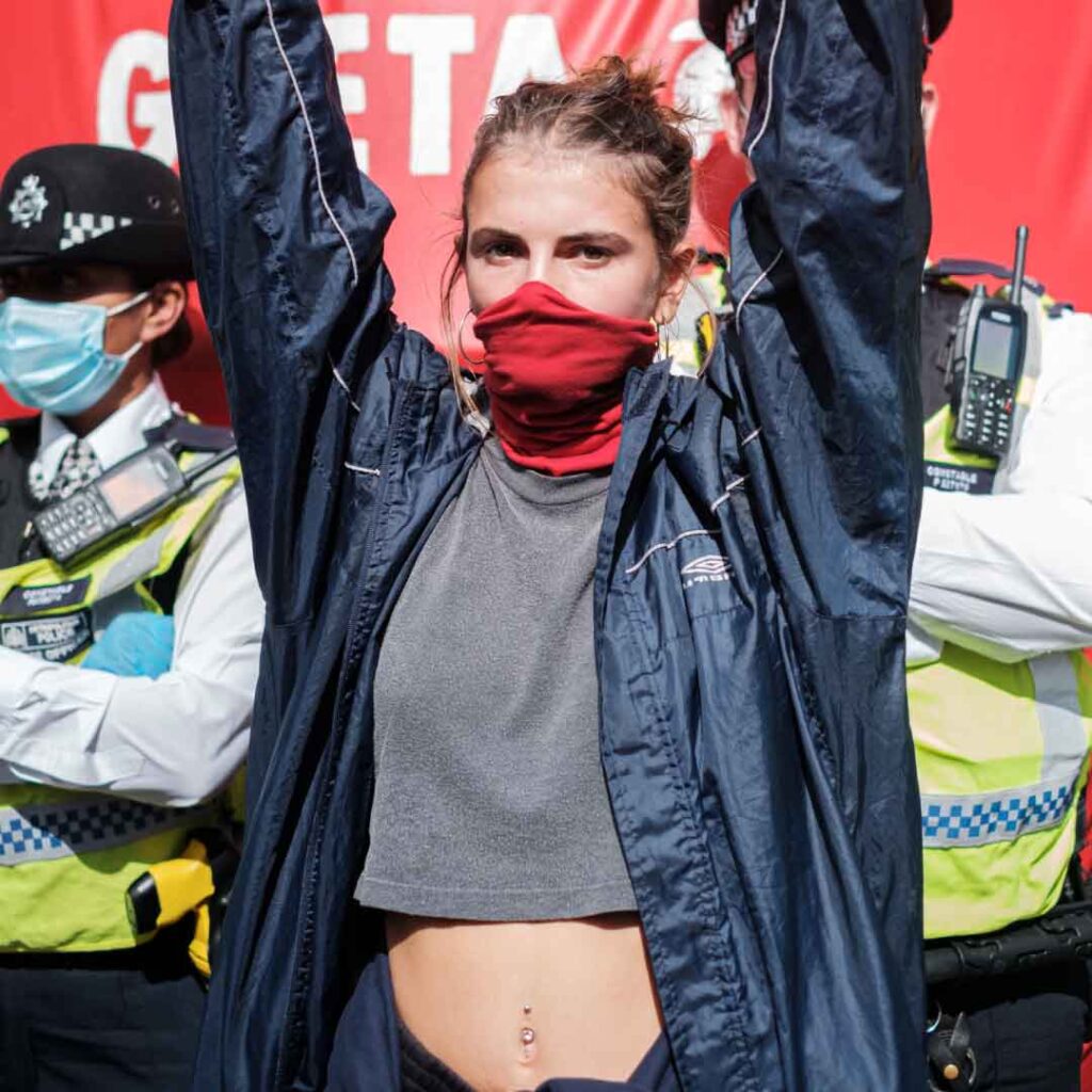 Photo of a young woman with a red bandana covering her mouth and her hands in the air. Two police officers are standing with arms folded behind her. Credit Ehimetalor Akhere Unuabona.