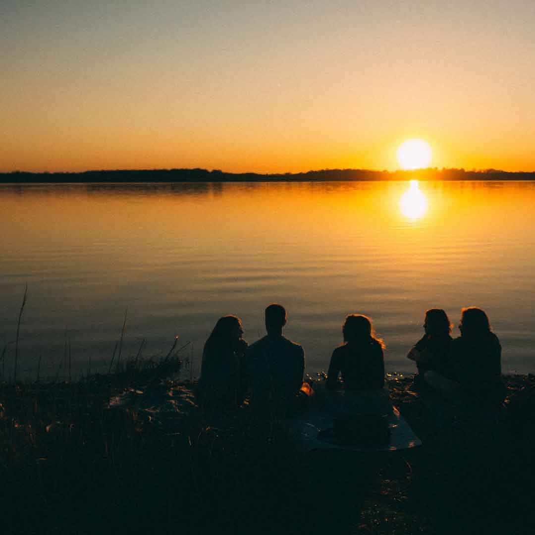 Photo of several young people sitting with a lake in front of them and the sun setting in the distance. Photo by Tobias Tullius on Unsplash
