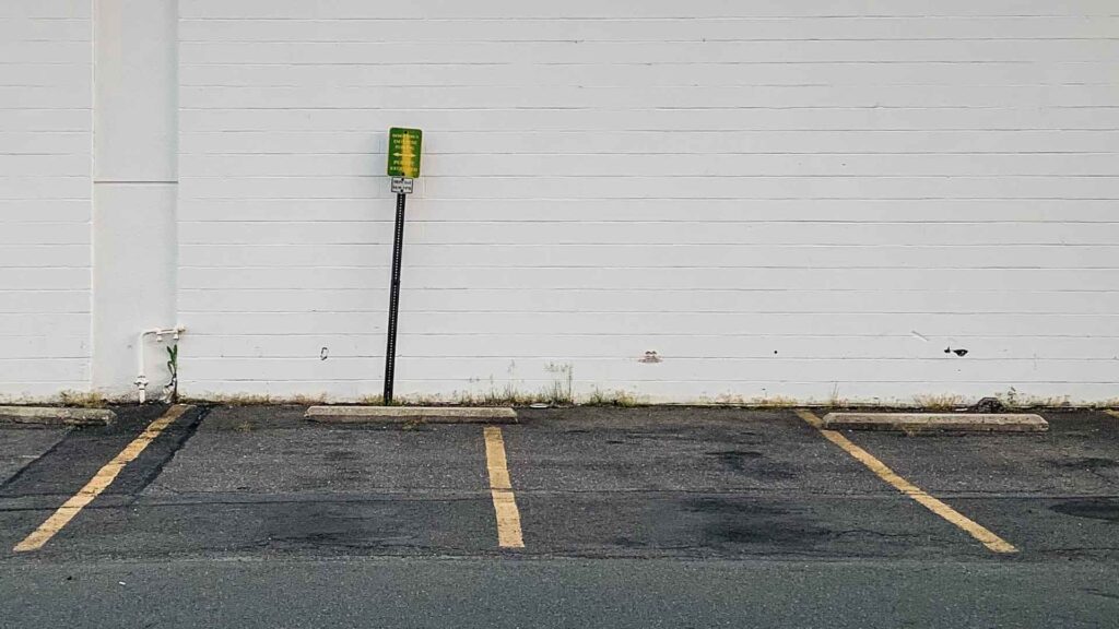 Photo of a parking lot, with yellow lot dividing lines stretching toward the viewing and a white building wall behind. Photo by Chris Mok || @cr.mok on Unsplash.