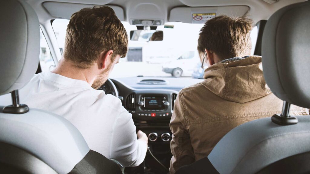 Photo of two people in the front of a car. One is in the driver's seat and the other in the passenger's seat. Both are facing away from the camera, which seems to be in the back seat. Both people appear to be men. Photo by David Emrich on Unsplash.