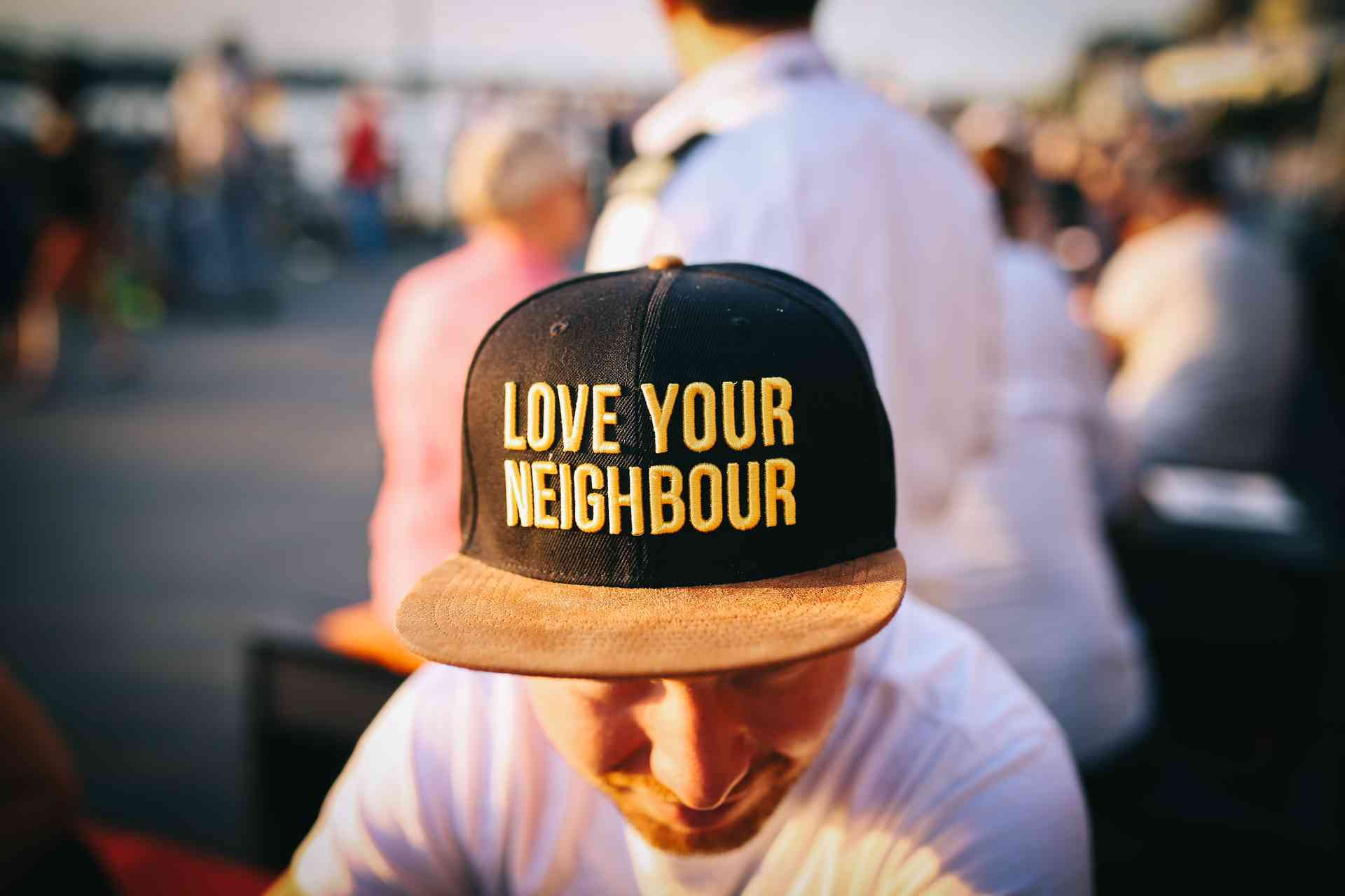 Photo of a man wearing a baseball cap, with the cap in focus and nearer the viewer. The cap has the words "Love your neighbour" on it. Photo by Nina Strehl on Unsplash