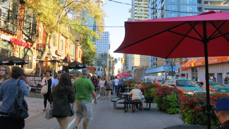 Photo of John St., Toronto, showing a street with many people walking on it, as well as people sitting on chairs at tables on the street. The widewalk has been extended to enhance walkability. Credit City of Toronto.