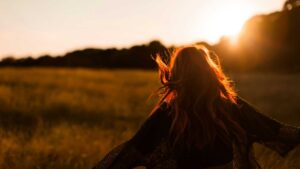 A person with long, flowing hair is shown from behind, standing in a sunlit field during sunset. The warm golden light creates a serene and peaceful atmosphere, with the silhouette of trees in the background. The person's hair catches the sunlight, highlighting its texture and color. The image evokes a sense of freedom and connection with nature. Photo by Sasha Freemind on Unsplash.