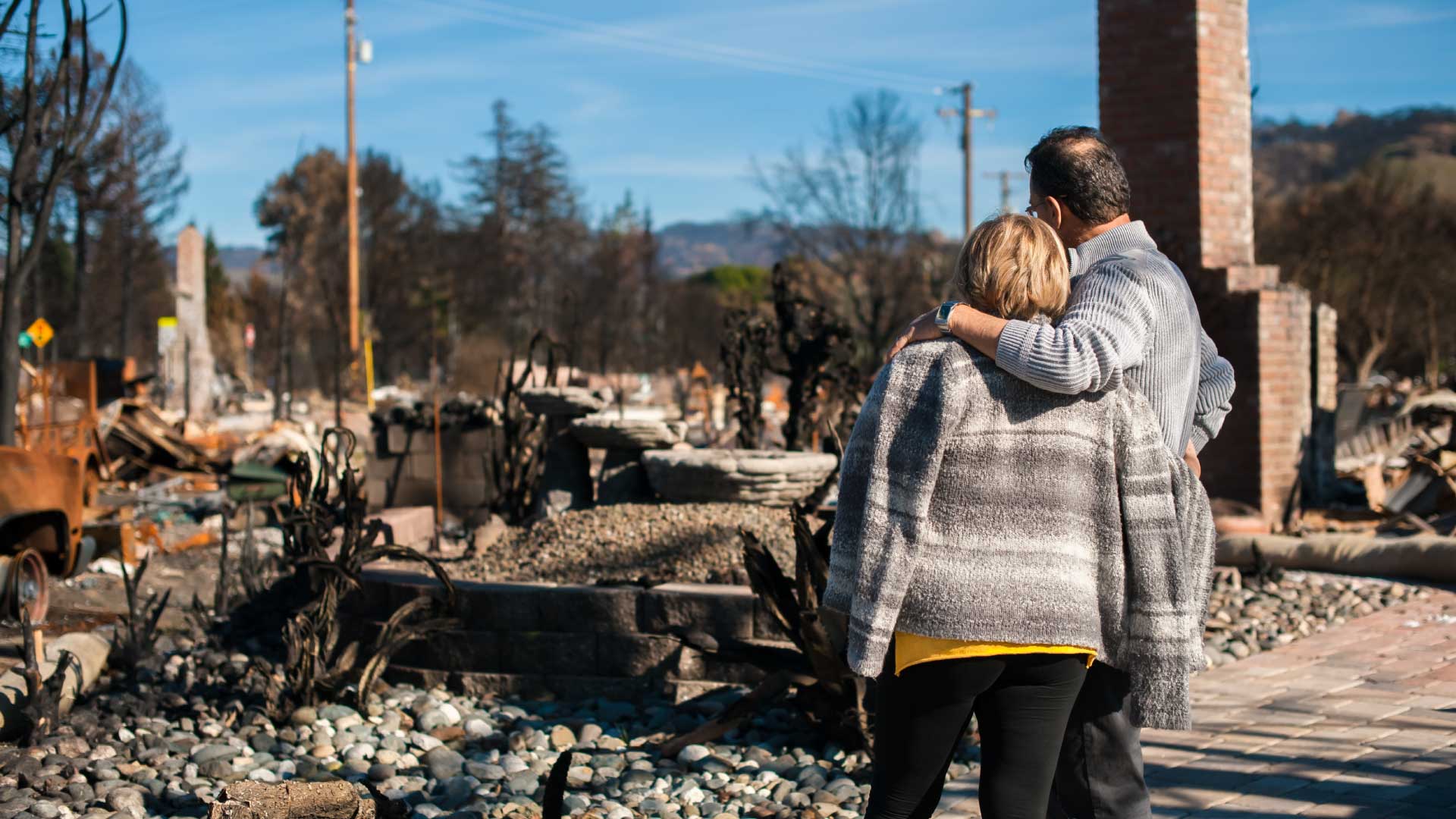 Photo of a couple, arms around each other, looking away from the camera at the ruins of a burned out house.