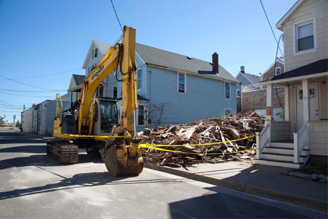 Photo showing damage from a natural disaster. A demolished house is shown with caution tape around the debris. An excavator is shown in front of the debris.